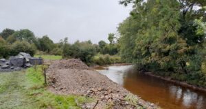 River to the right beside trees and to the left of the river there are piles of gravel that have been removed from the riverbed